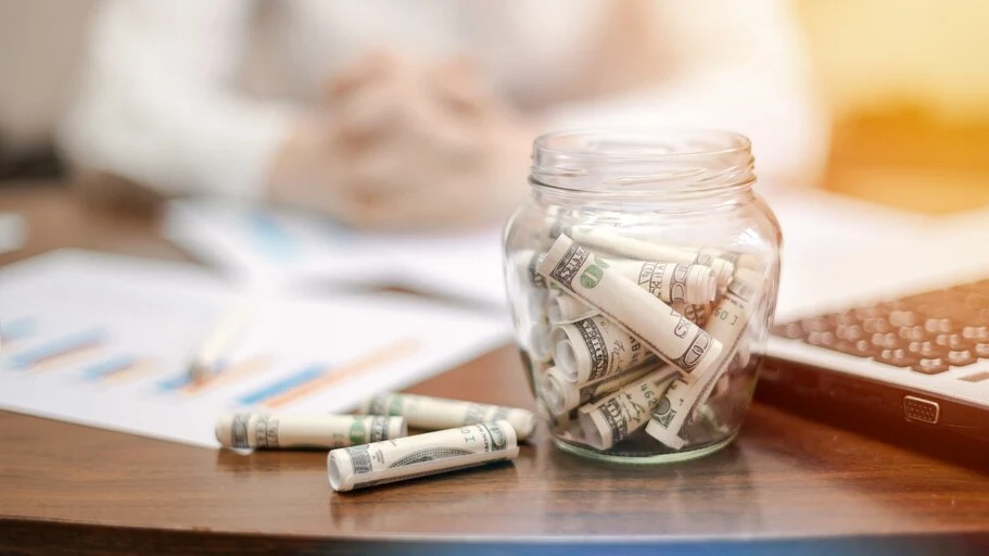 jar-with-rolled-banknotes-table-laptop-papers-woman-background